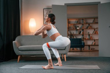 Çömeliyor. Beyaz spor giysili zayıf vücutlu genç kadın içeride..