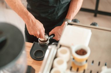 Kahve makinesiyle çalışmakla meşgul. Beyaz gömlekli ve siyah önlüklü kafe çalışanı içeride..