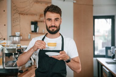 Ayakta ve bardağı tutarken. Beyaz gömlekli ve siyah önlüklü kafe çalışanı içeride..