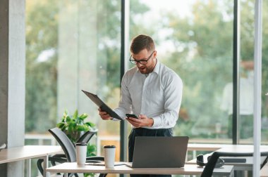 Kafam karıştı. Belgeler ve telefonlar duruyor. Resmi kıyafetli adam modern ofiste çalışıyor..