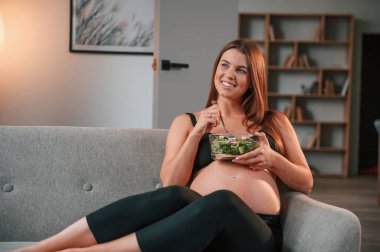 Kanepede oturup salata yemek. Güzel hamile kadın evde..