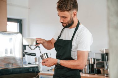 Ayakta ve kahve doldururken. Beyaz gömlekli ve siyah önlüklü kafe çalışanı içeride..