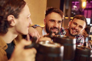 Masanın yanında oturuyor. Üç arkadaş barda bira içip iyi vakit geçiriyorlar..