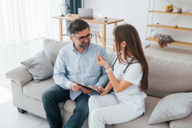 Kadın doktor koltukta olgun bir müşteriyle oturuyor. Ofiste beyaz önlüklü profesyonel bir sağlık görevlisi var..