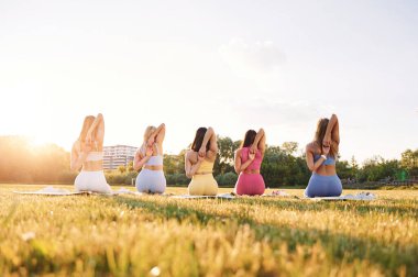 Lotus pozisyonunda oturuyor. Bir grup kadın sahada birlikte spor yapıyorlar..