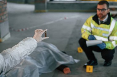 Bir kadın mermi kovanı tutuyor. Dedektifler cesedin yanındaki suç mahallinde kanıt topluyorlar..