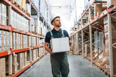 Elinde beyaz bir kutuyla yürüyorsun. Üniformalı eleman gündüz vakti depoda çalışıyor..