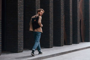 Sakallı, haki renkli ceketli ve kot pantolonlu şık bir adam binanın yanında..