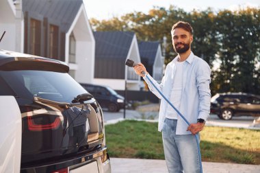 Ekoloji kavramı. Şarj kablosuyla duruyorum. Genç şık adam, gündüz vakti elektrikli arabayla geziyor..