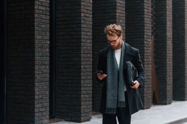 Akıllı telefonu tutuyorsun. Sakallı ve gözlüklü şık adam binanın yanında..