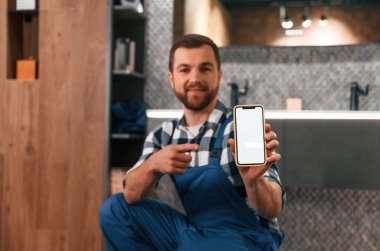Akıllı telefonu tutuyorsun. Mavi üniformalı tesisatçı banyoda çalışıyor..