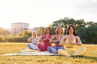 Lotus pozisyonunda oturuyor. Bir grup kadın sahada birlikte spor yapıyorlar..