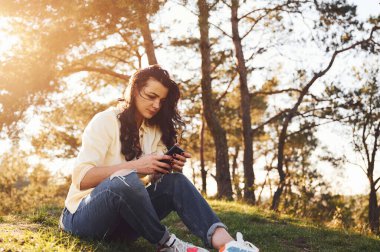 Akıllı telefon kullanıyor. Güzel bir kadın gündüz vakti ormanda çimlerin üzerinde oturuyor..