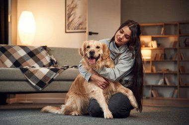 Oturuyor ve gülümsüyor. Kadın Golden Retriever köpeğiyle evde..
