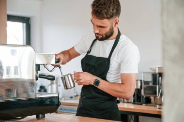 Ayakta ve kahve doldururken. Beyaz gömlekli ve siyah önlüklü kafe çalışanı içeride..