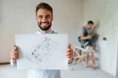 Oda planı tutuyor ve gülümsüyor. İki adam dairede birlikte tamirat yapıyor..