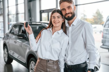 Ellerinde anahtarlarla dikiliyorlar. Mutlu sahipler. Beyaz elbiseli bir adam araba galerisinde birlikte..