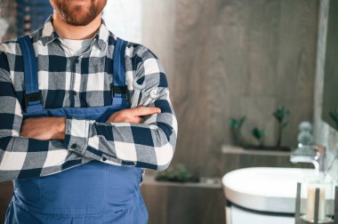 Kollarını kavuşturmuş duruyorsun. Mavi üniformalı tesisatçı banyoda çalışıyor..