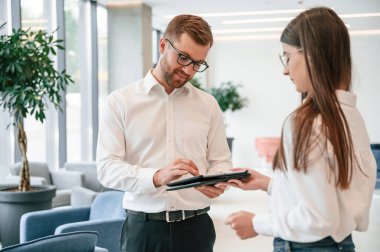 Elinde tabletle. Resmi giyinmiş kadın ve erkek içeride birlikte çalışıyorlar..