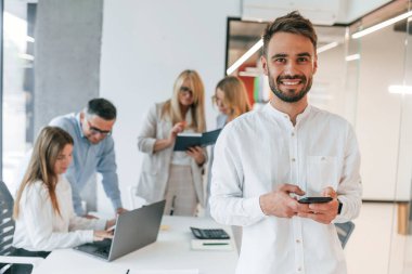 Kameraya poz veren güzel, neşeli bir adam. Profesyonel iş adamlarından oluşan bir grup ofiste..