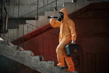 El feneri ve çantayla merdivenlerde. Kıyamet sonrası binanın harabelerinde kimyasal koruma kıyafeti giymiş bir adam..