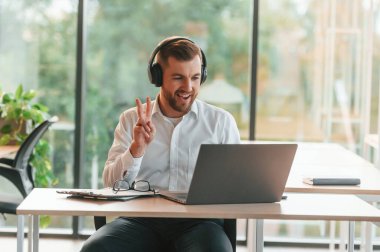 Laptop, kulaklık, olumlu bakış. Resmi kıyafetli adam modern ofiste çalışıyor..