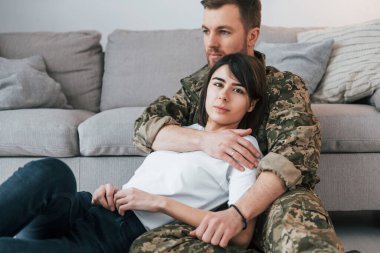 Yerde oturup, kadını kucaklamak gibi. Üniformalı asker evde karısıyla birlikte..