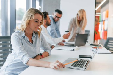 Olgun bir kadın dizüstü bilgisayarla masada oturuyor. Profesyonel iş adamlarından oluşan bir grup ofiste..
