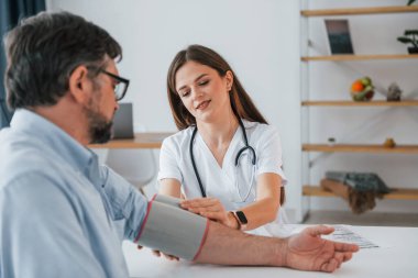 Kadın doktor adamın kan basıncını ölçüyor. Ofiste beyaz önlüklü profesyonel bir sağlık görevlisi var..