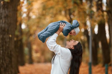 Güzel anne küçük oğlunu elinde tutuyor. Sonbahar ormanında dikilirken.