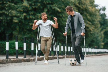 Koltuk değnekli iki adam yolda. Bir futbol topuyla.