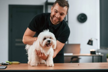 Adam gülümsüyor. Groomer içeride Maltalı köpekle birlikte. Siyah üniformalı adam..