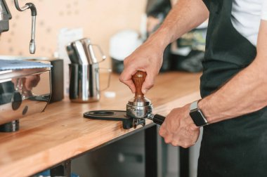 Kahve makinesiyle çalışmakla meşgul. Beyaz gömlekli ve siyah önlüklü kafe çalışanı içeride..