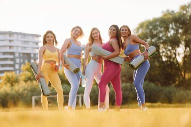 Gülümsüyor ve ayakta duruyor. Bir grup kadın sahada birlikte spor yapıyorlar..