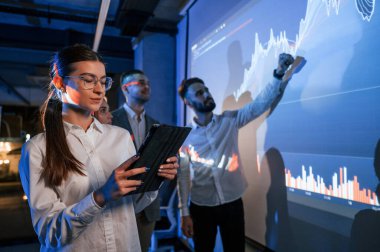 Kadın tabletle ayakta duruyor. Erkek lider çalışanlarıyla konuşuyor, projektörle ilgili planı ofislerinde gösteriyor..