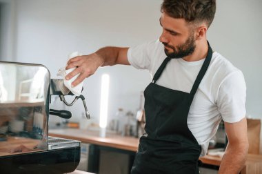 Kahve makinesini temizliyorum. Beyaz gömlekli ve siyah önlüklü kafe çalışanı içeride..