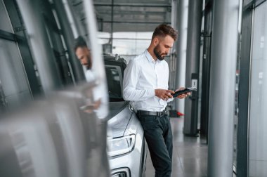 Elinde siyah bir tablet var. Beyaz elbiseli genç adam araba galerisinde..