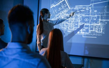 İşçilerle konuşan kadın lider, borsa şirketinin projektörüyle ilgili planı gösteriyor..