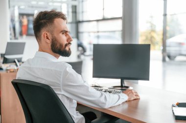 Masanın yanında oturuyor. Beyaz elbiseli genç adam araba galerisinde..