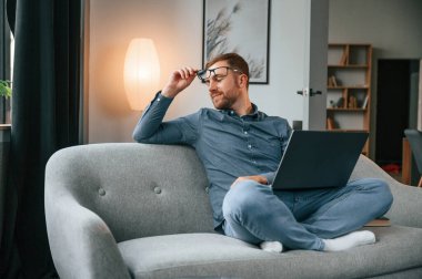 Oturup yan tarafa bakıyor. Erkek serbest çalışan evde dizüstü bilgisayar kullanıyor. Konforlu daire.