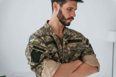 Kollar çapraz. Üniformalı asker içeride beyaz duvara karşı duruyor..