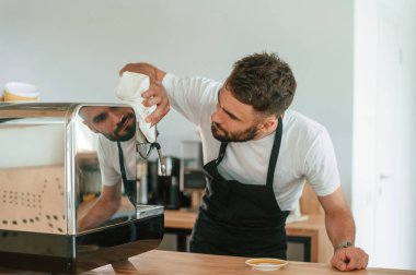 Kahve makinesini temizlemekle meşgul. Beyaz gömlekli ve siyah önlüklü kafe çalışanı içeride..