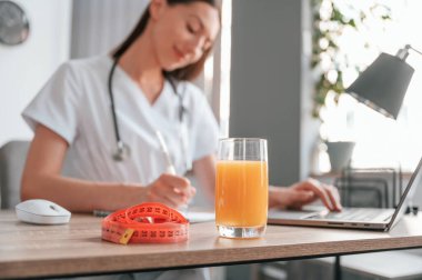 Dizüstü bilgisayarla çalışıyor. Elma suyu ve ölçü uzunluğu masanın üzerinde. Genç, güzel bayan doktor..