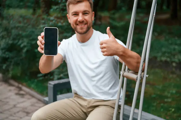 stock image Holding and showing smartphone. Man with crutches is in the park outdoors. Having leg injury.