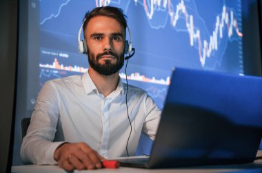 Dizüstü bilgisayarın yanında oturuyor. Ofiste borsa simsarı ona karşı büyük ekranla işlem yapıyor..