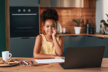 Çizim için dizüstü bilgisayar ve kağıtla. Gündüz vakti evde rahat giyinen sevimli siyah kız var..