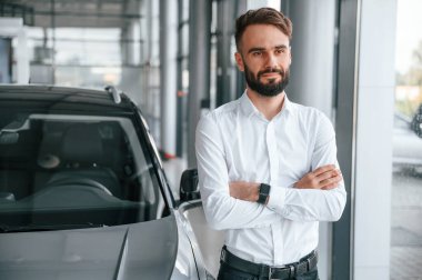 Kollarını kavuşturmuş duruyorsun. Beyaz elbiseli genç adam araba galerisinde..