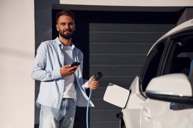 Akıllı telefon ve şarj kablosu ellerinde. Genç şık adam, gündüz vakti elektrikli arabayla geziyor..