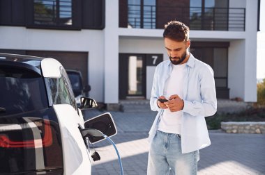Şarj edilmeyi bekliyor, akıllı telefonu tutuyor. Genç şık adam, gündüz vakti elektrikli arabayla geziyor..