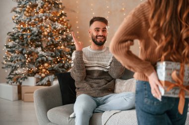 Kadın sürpriz yapıyor, elinde hediye kutusu var. Güzel genç çift yeni yılı evde kutluyor..